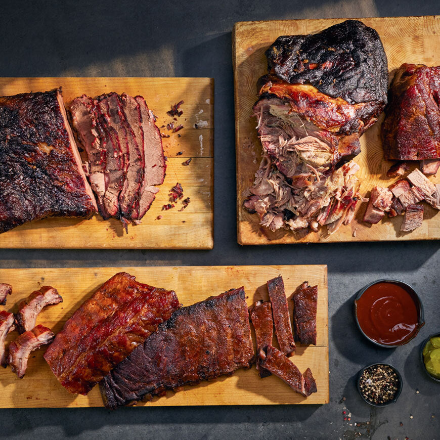 Three wooden cutting boards with sliced smoked meats and a small bowl of sauce on a dark surface.