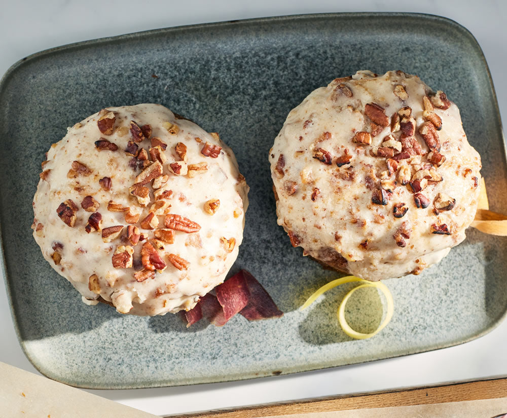 Two carrot cakes with frosting and nut topping on a textured plate with a lemon wedge.